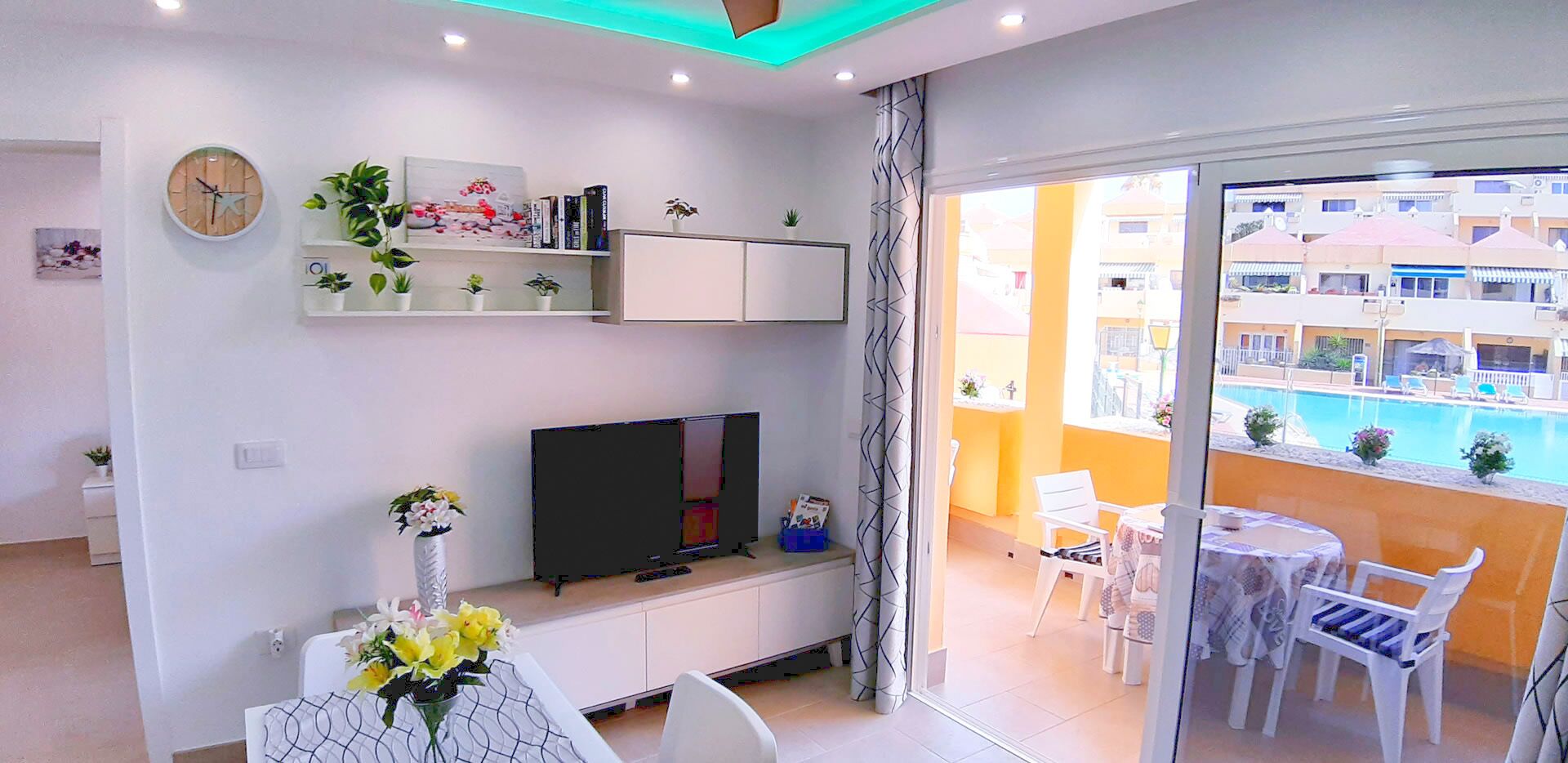 A modern living room with a flat-screen TV, a dining table with a vase of flowers, and a balcony with a pool view.