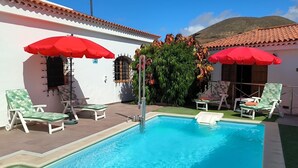 A swimming pool with lounge chairs and red umbrellas, surrounded by a white building with a tiled roof.