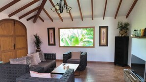 A spacious living room with a high ceiling, wooden beams, a large window with a view of greenery, a television on a stand, and a black cabinet.