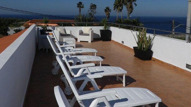 A rooftop terrace with white lounge chairs and potted plants, overlooking a coastal view.