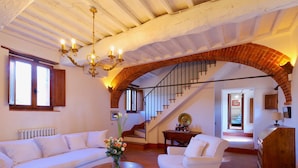 A spacious living room with a white sofa, a white armchair, a coffee table, and a chandelier. There is a staircase with a wooden railing leading to another room. - Chiusarella Arceno Rentals Club Chianti Villa exclusive Pool Concierge freeWiFi (CASTELNUOVO BERARDENGA)