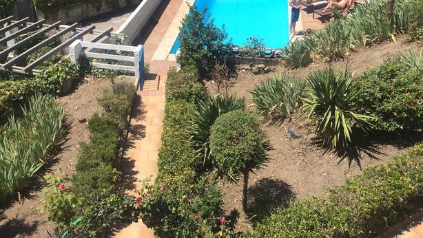 A rectangular swimming pool surrounded by a white fence, with a paved deck and a few people relaxing nearby.