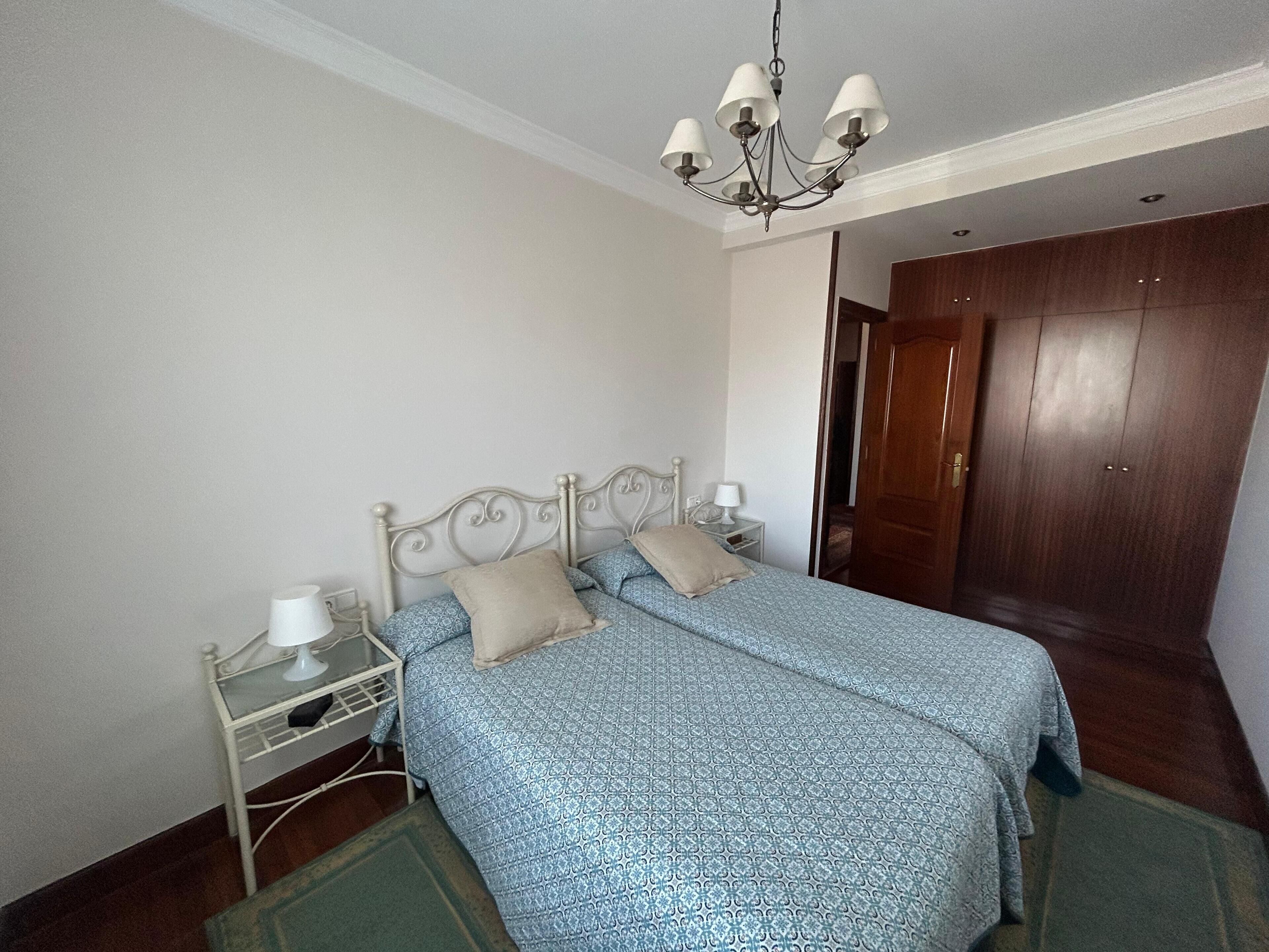 Een slaapkamer met een tweepersoonsbed, een nachtkastje, een lamp, een houten garderobe en een kroonluchter.