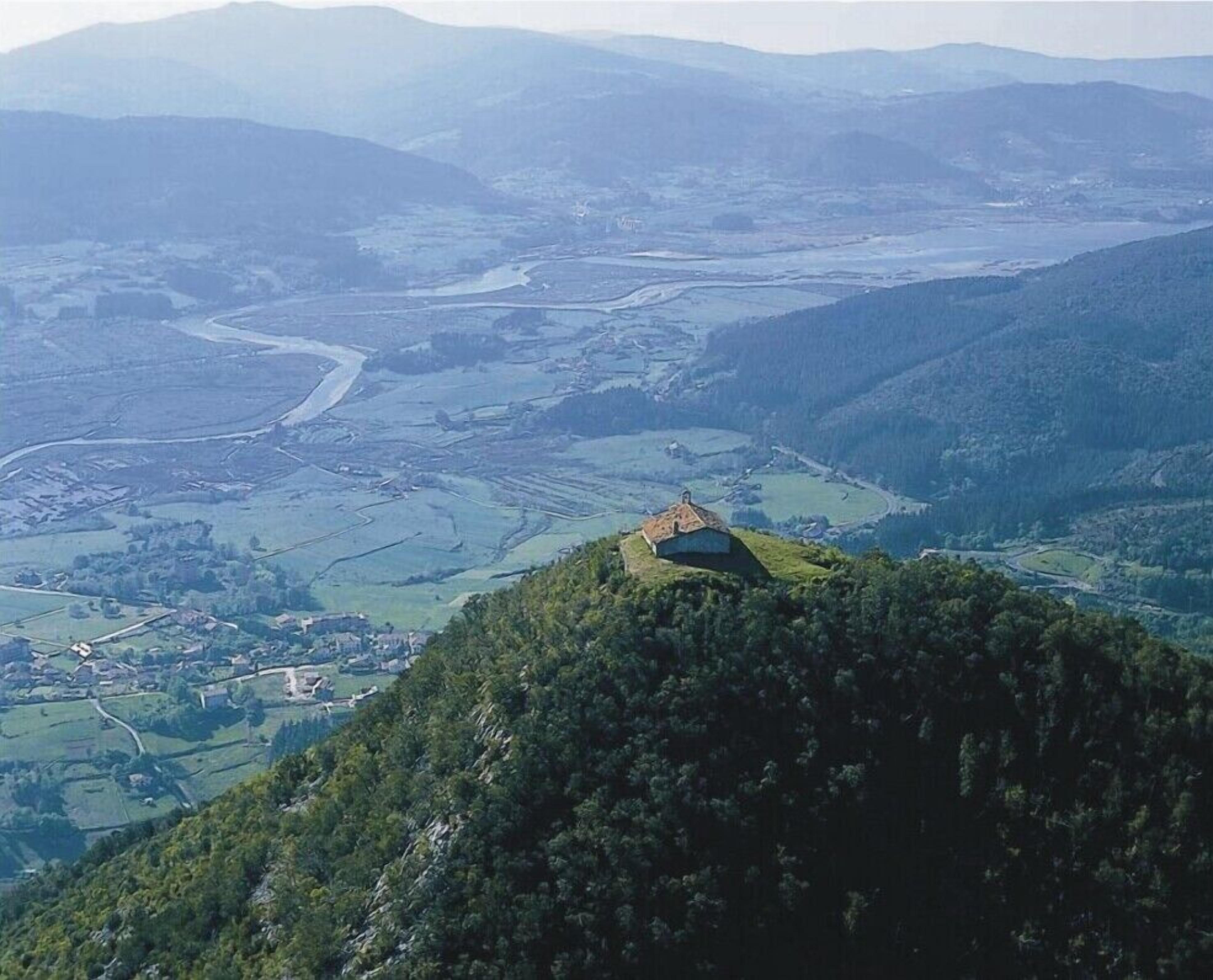 A panoramic view of a valley with a prominent hill featuring a small building, surrounded by dense forests and distant mountains.