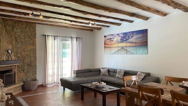 A cozy living room with a stone fireplace, a large gray sectional sofa, a wooden dining table with chairs, and a painting of a sunset on the wall. - Lagoa Albufeira House - with private swimming pool (Sesimbra)