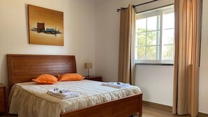 A neatly made bed with a wooden headboard, a bedside table with a lamp, and a window with curtains. - Lagoa Albufeira House - with private swimming pool (Sesimbra)