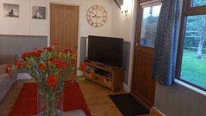 Ein Wohnzimmer mit Holzboden, einem Fernseher auf einem Holztisch, einem roten Teppich, einer Vase mit roten Blumen und einer Uhr an der Wand.