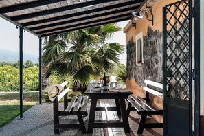 Een terras buiten met een houten tafel en banken, omgeven door een tuin met palmbomen.