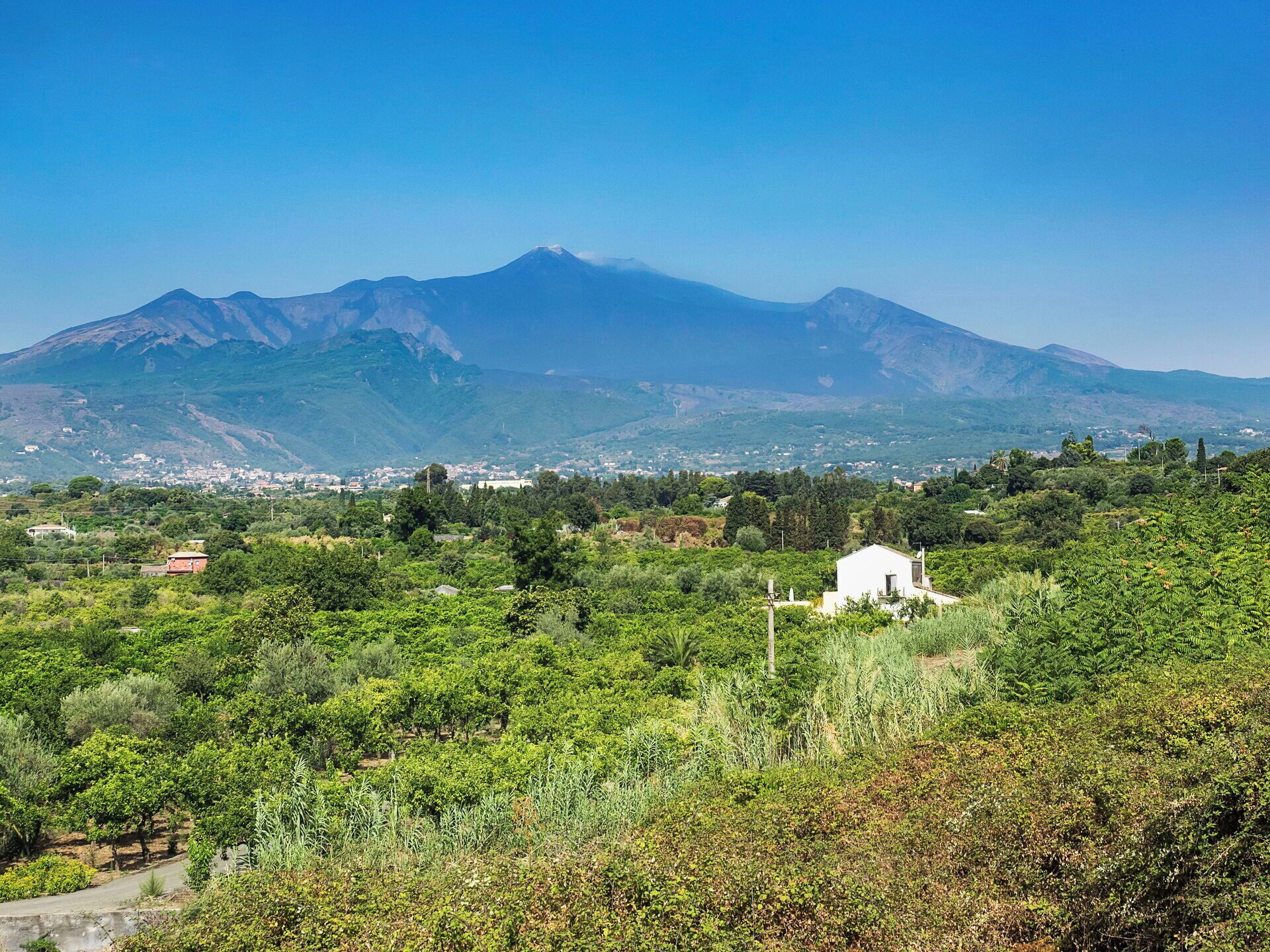 Paisaje montañoso con un edificio blanco en primer plano, rodeado de vegetación exuberante y un pueblo al fondo.