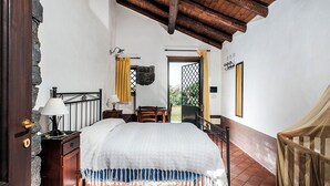 A bedroom with a bed, a nightstand, a lamp, a chair, a wooden desk, a window with curtains, and a door leading outside. - Country-house with pool and Etna and sea views (Acireale)
