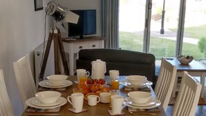 Une table à manger dressée avec de l’assiette blanche, des tasses et des bols, accompagnée d’un saladier de fruits et d’un jus d’orange.