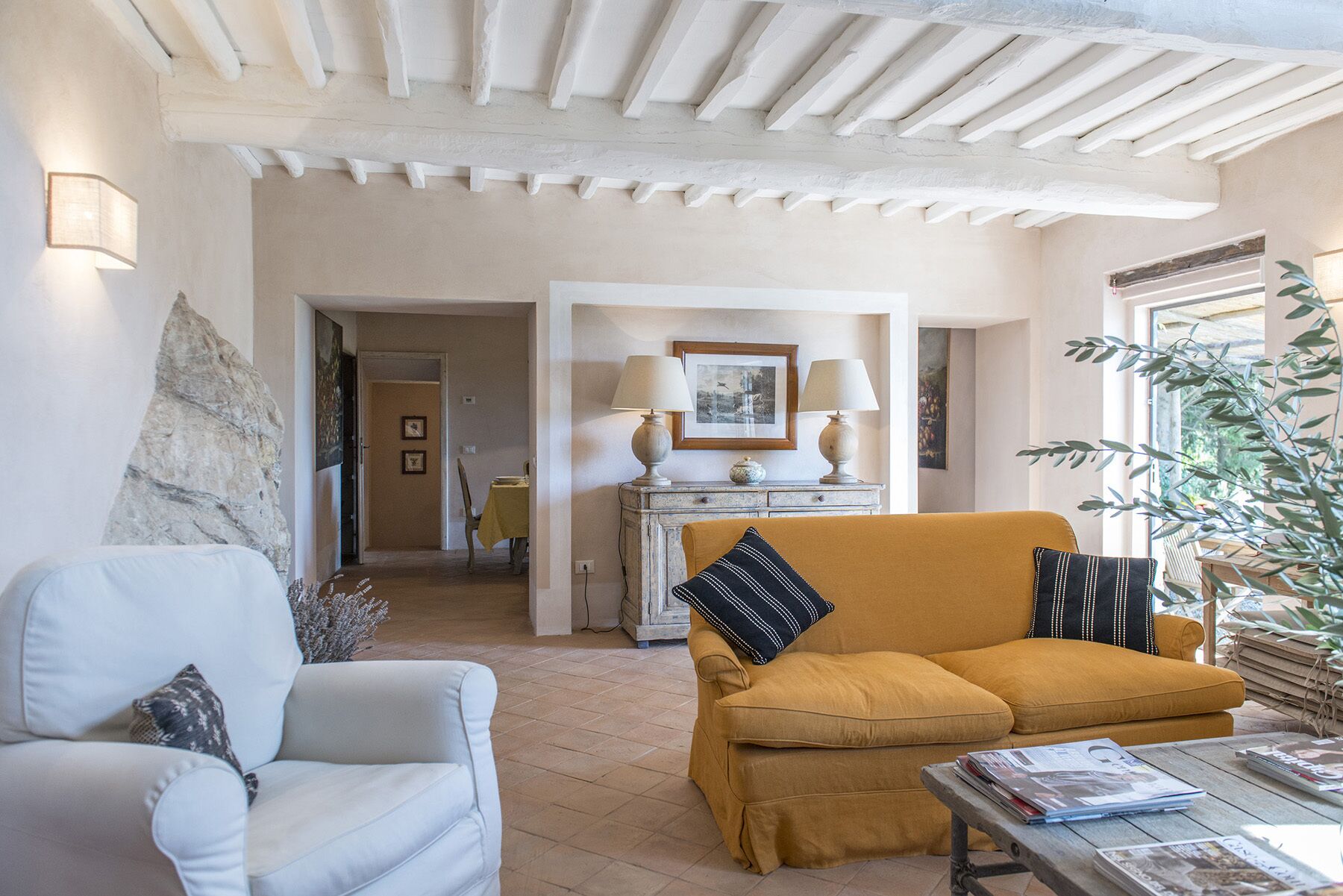 Un salon avec un canapé blanc, un canapé de couleur moutarde et une table basse en bois sur laquelle sont posés des magazines. Il y a un tableau au mur et une lampe sur une table d’appoint.