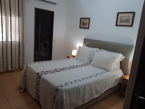 Une chambre à coucher avec un lit, une table de chevet, une lampe, une porte et deux tableaux encadrés au mur.