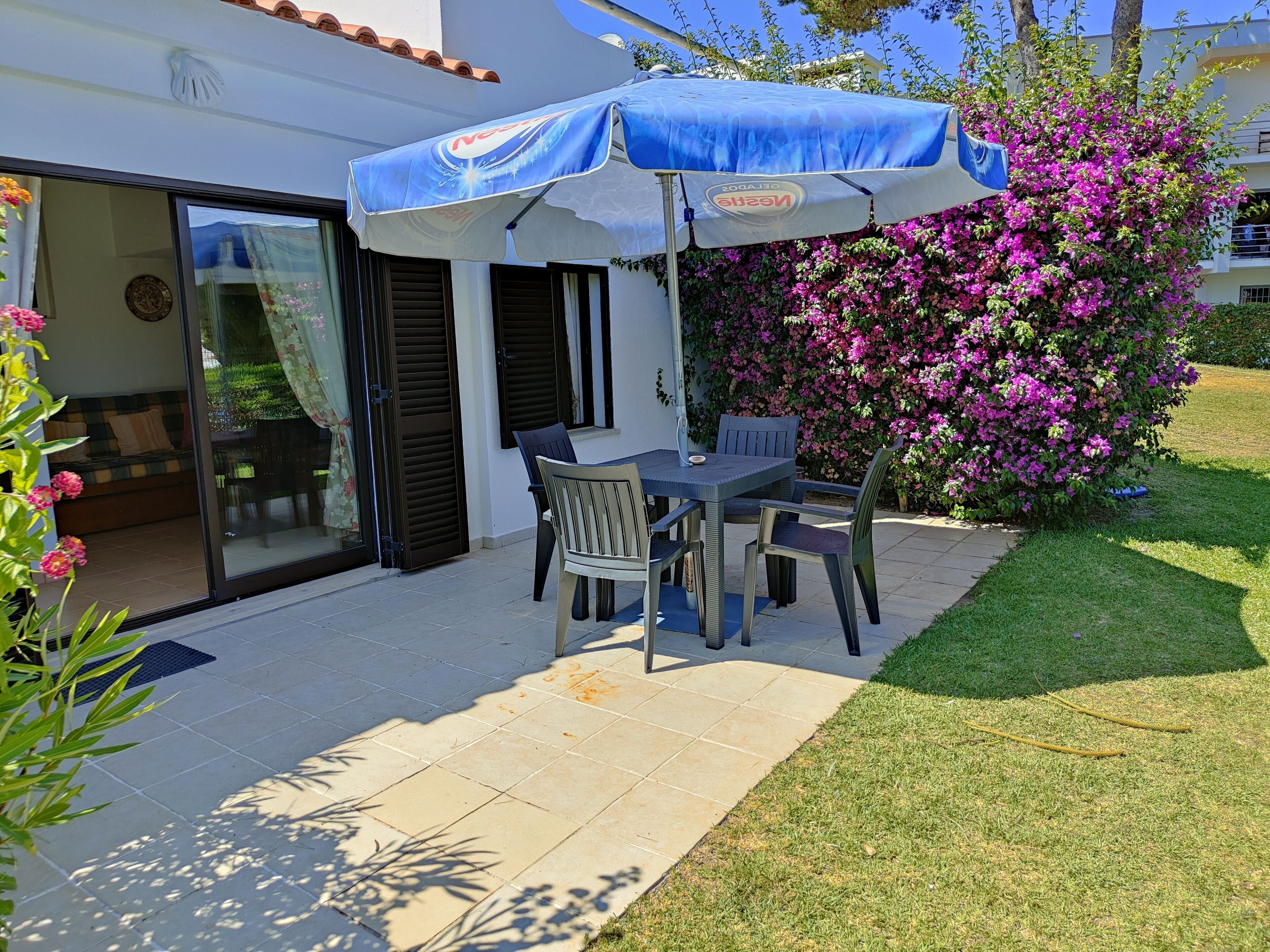 An outdoor patio with a table and chairs, a blue umbrella, and a garden with pink flowers.