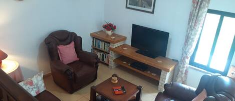 A living room with a brown leather sofa, a wooden coffee table, a television on a wooden stand, a bookshelf, and a window with curtains.
