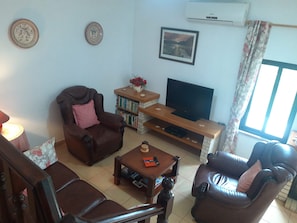 A living room with a brown leather sofa, a wooden coffee table, a television on a wooden stand, a bookshelf, and a window with curtains.