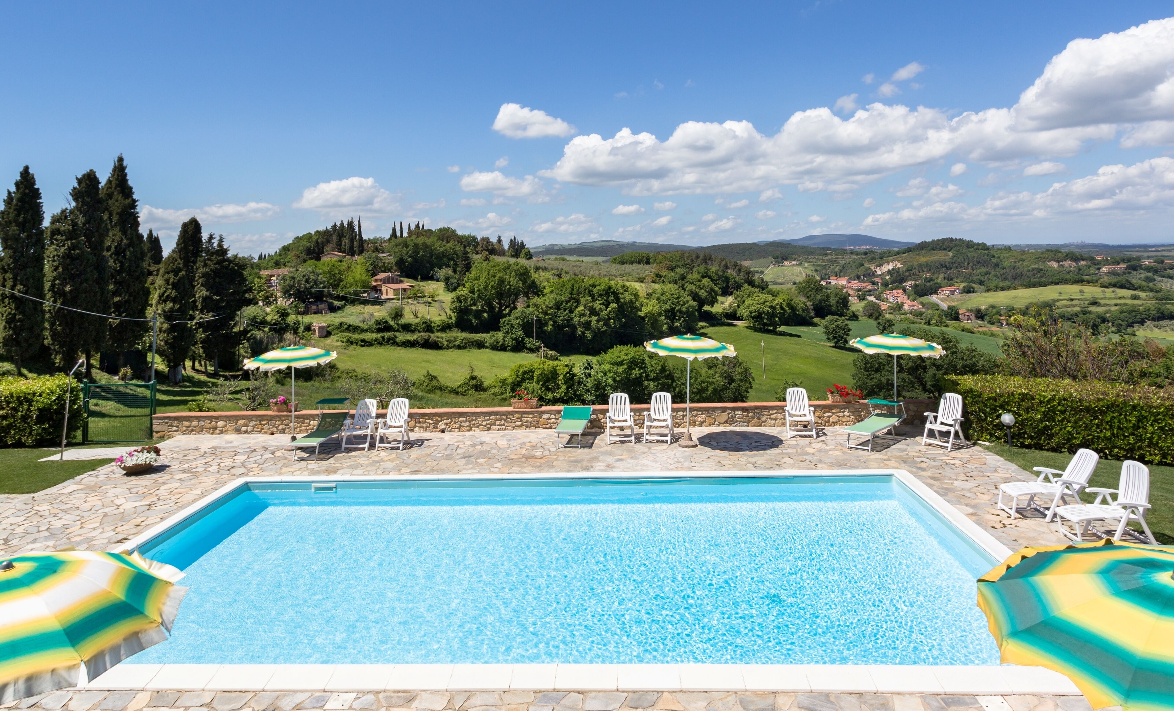 Una piscina con sillas de descanso y sombrillas, rodeada por una terraza de piedra y una vista panorámica de colinas y árboles.