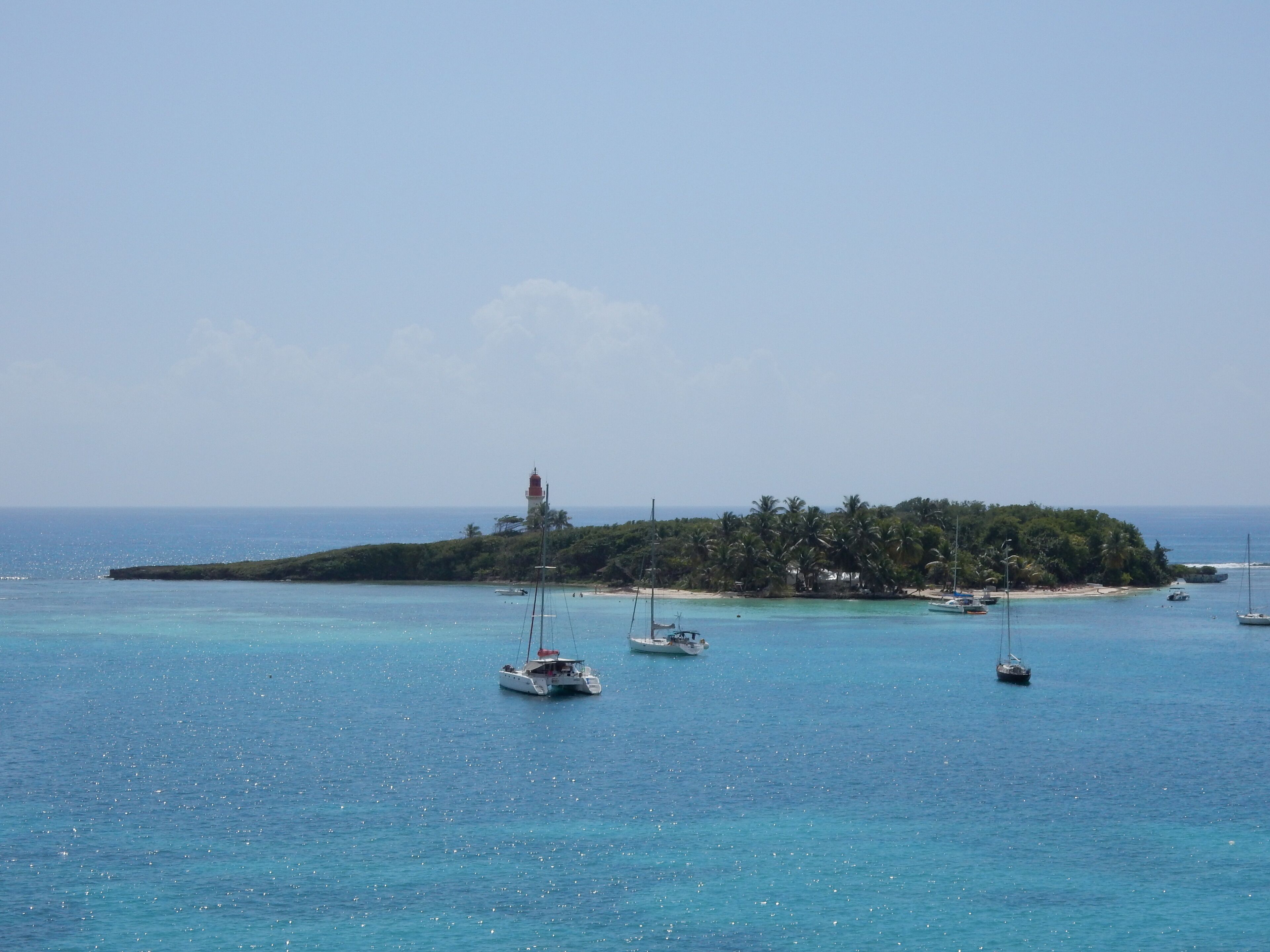A coastal scene with a small island featuring a lighthouse, surrounded by clear blue waters with several boats anchored.