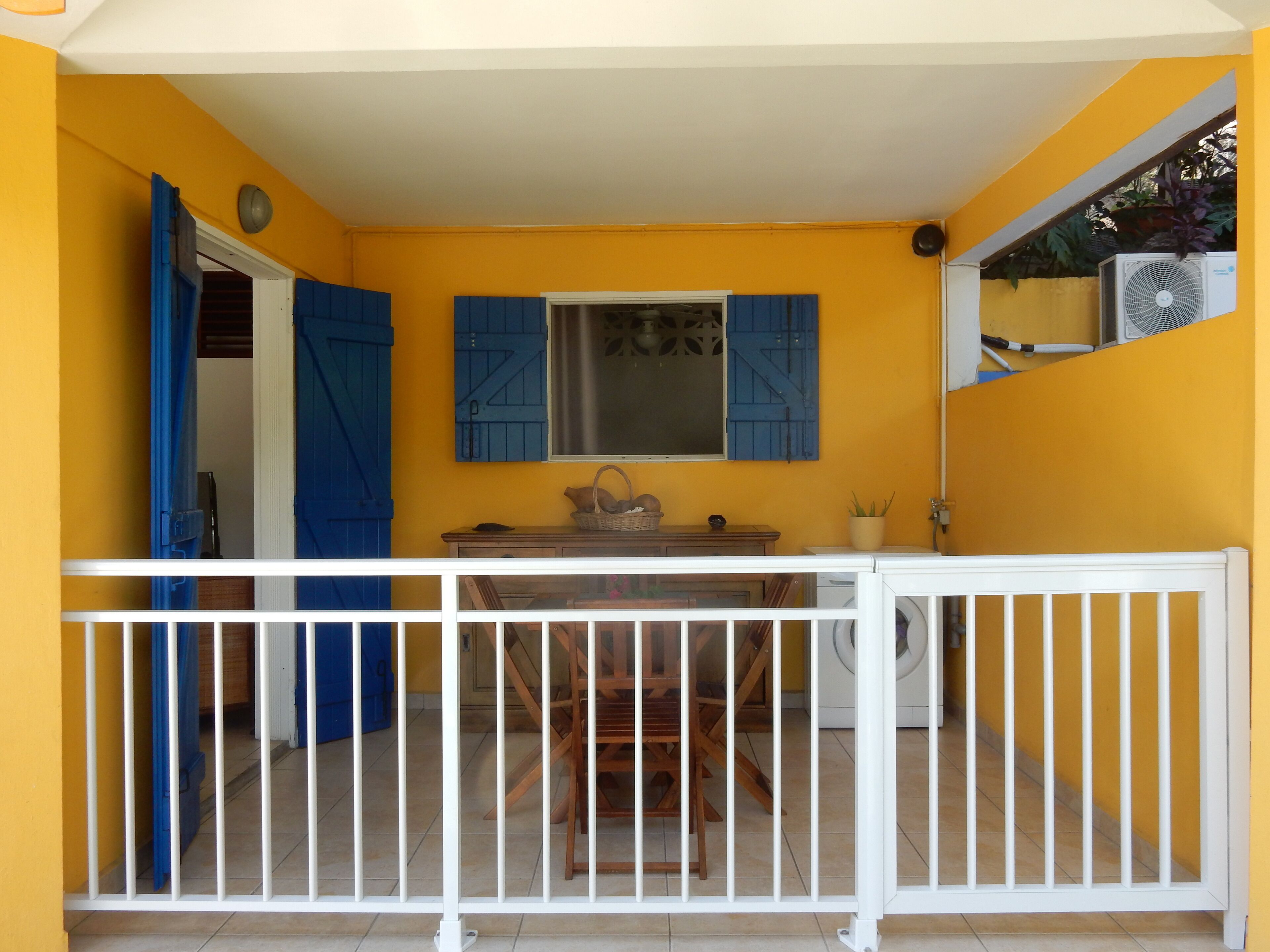 A small, enclosed balcony with a white railing, a wooden table, and a window with blue shutters.