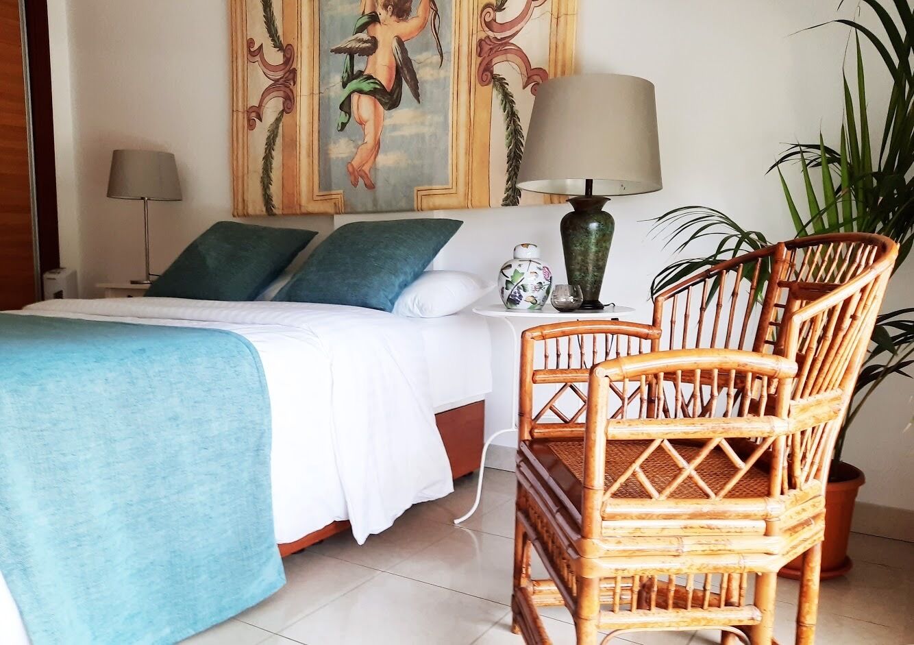 A neatly arranged bedroom with a bed, a bedside table, a lamp, a decorative vase, and a wicker chair.