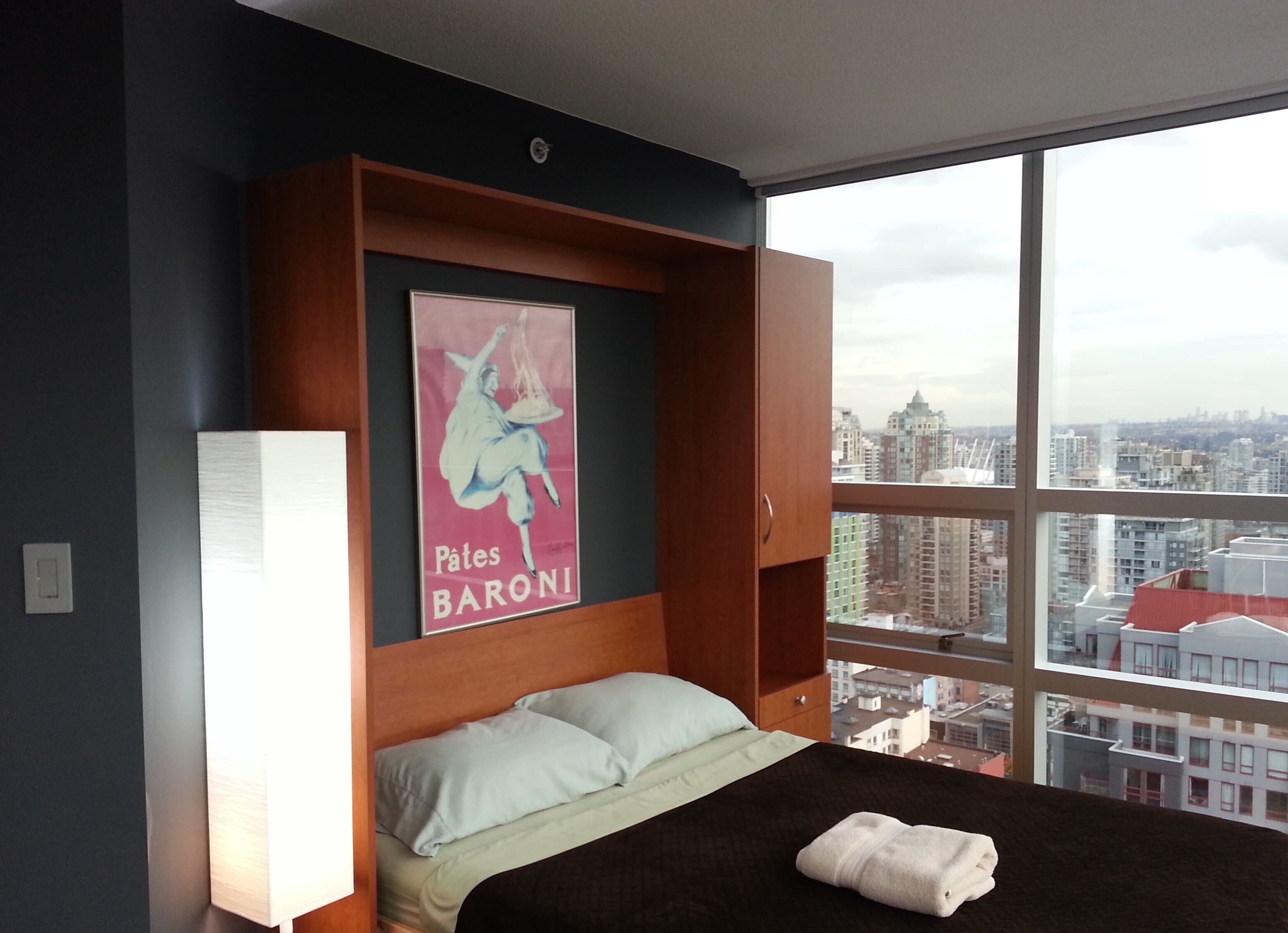 Habitación de hotel con una cama grande, una mesita de noche, una lámpara de pared y un cartel con la imagen de una mujer y el texto "Pates BARONI".