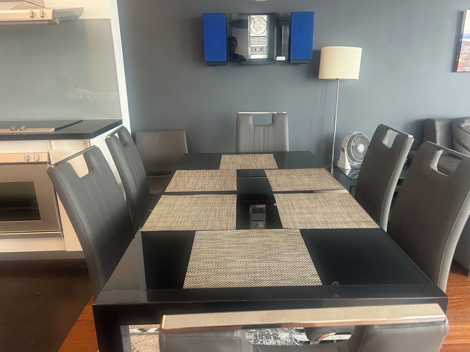 A modern dining area with a rectangular table, six chairs, a central air conditioning unit, a clock, and a lamp on the wall.