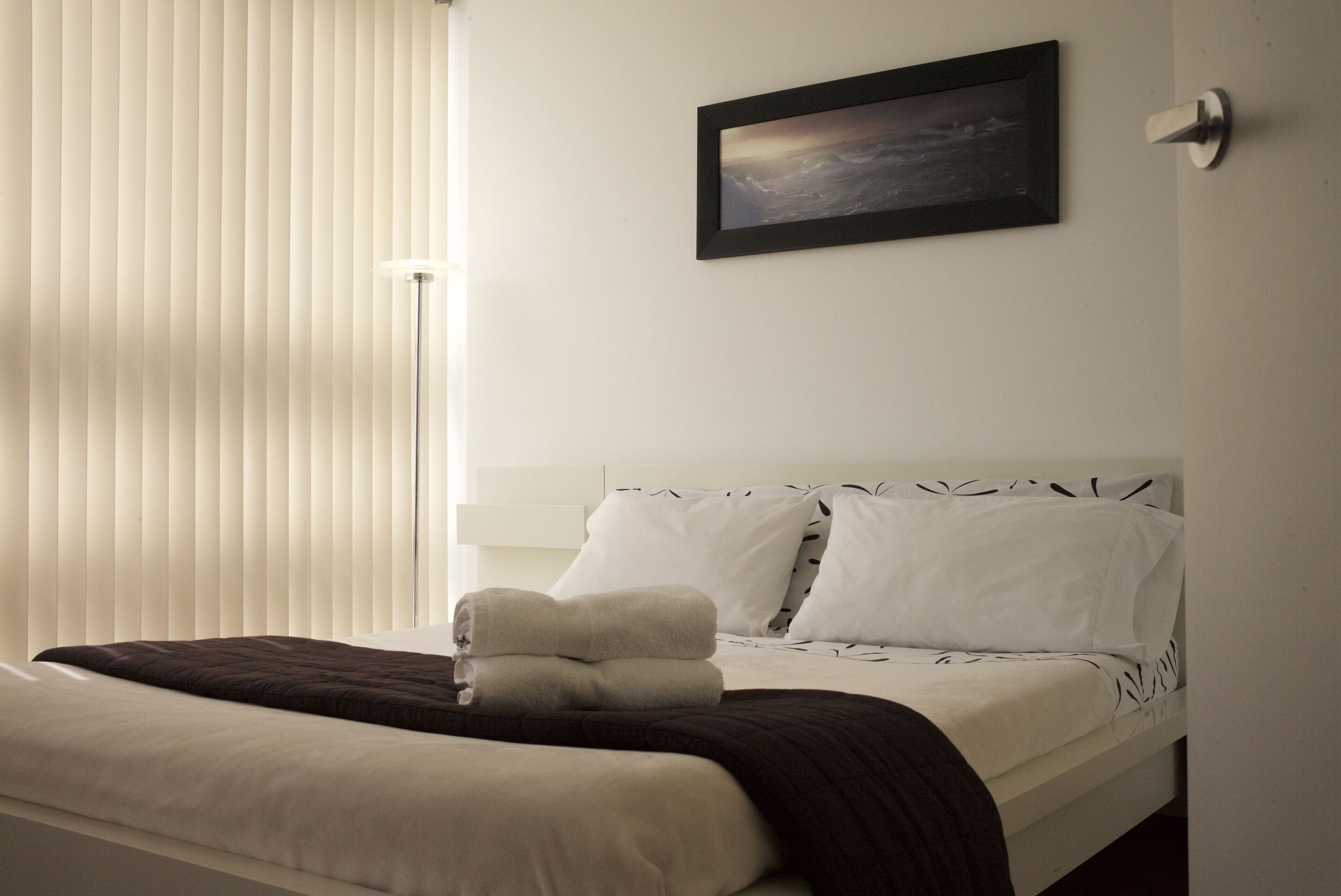 A neatly made bed with white and dark brown bedding, a framed picture on the wall, and a window with blinds.