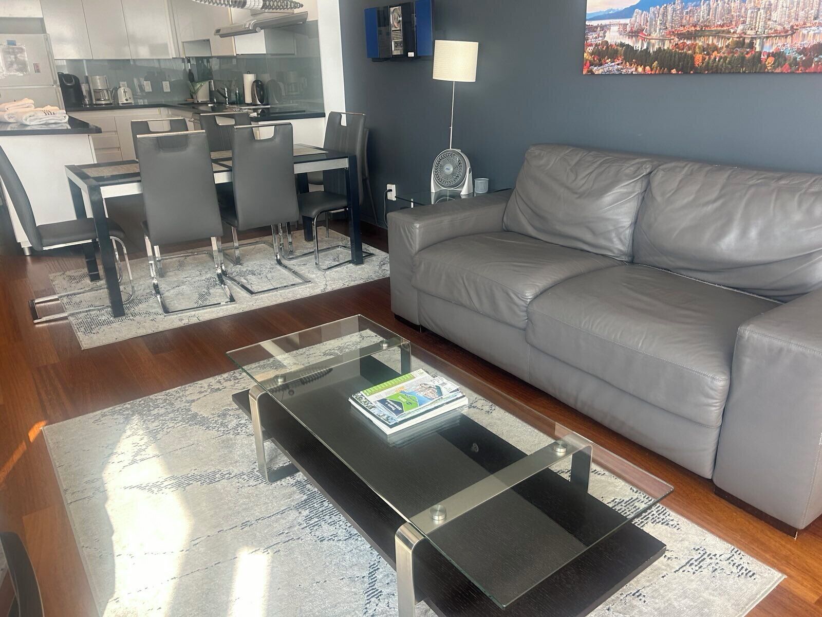 Un salon moderne avec un canapé gris, une table basse en verre et une cuisine en arrière-plan.