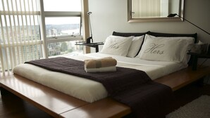 Ein ordentlich bezogenes Bett mit einer Sitzbank am Fußende, ein Nachttisch mit Lampe und ein Fenster mit Jalousien.