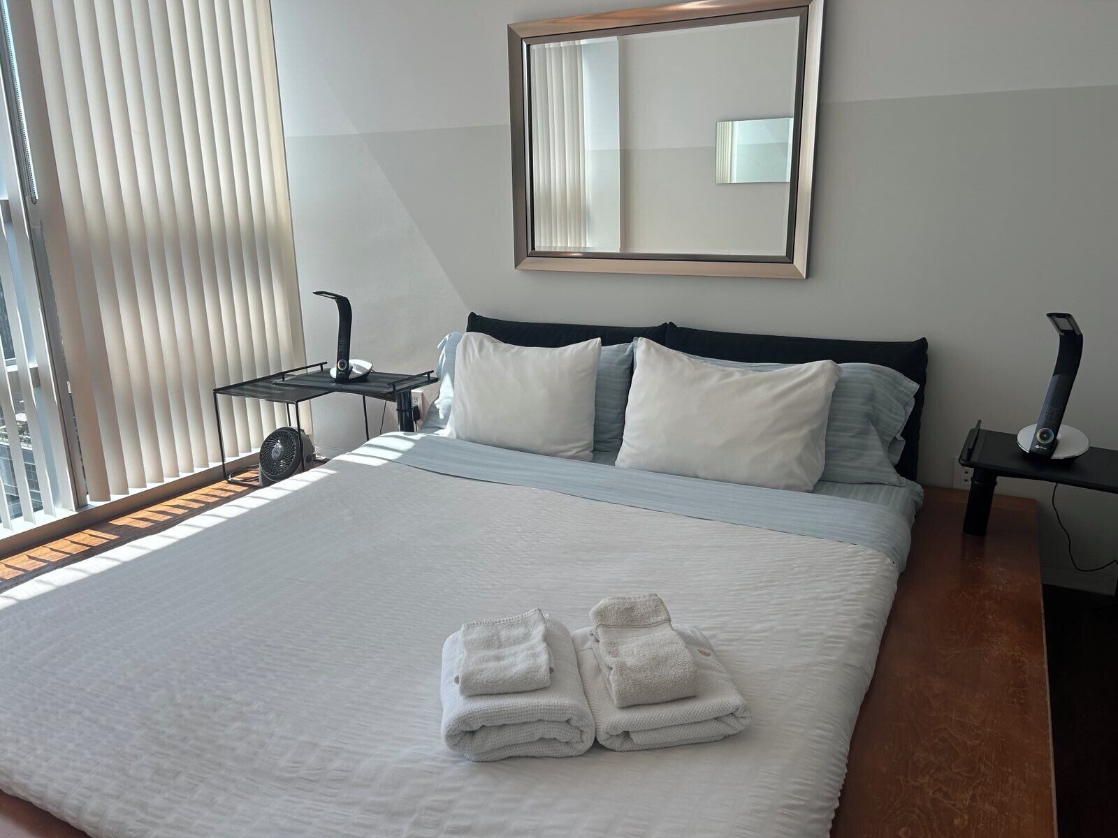 Una cama bien hecha con sábanas blancas y dos toallas dobladas, una mesita de noche con una lámpara y un espejo grande en la pared.