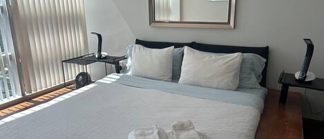 Una cama bien hecha con sábanas blancas y dos toallas dobladas, una mesita de noche con una lámpara y un espejo grande en la pared.