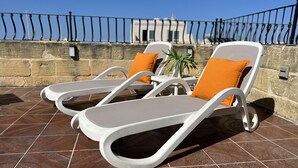 A terrace with two white lounge chairs, each with an orange cushion, and a small table with a potted plant.