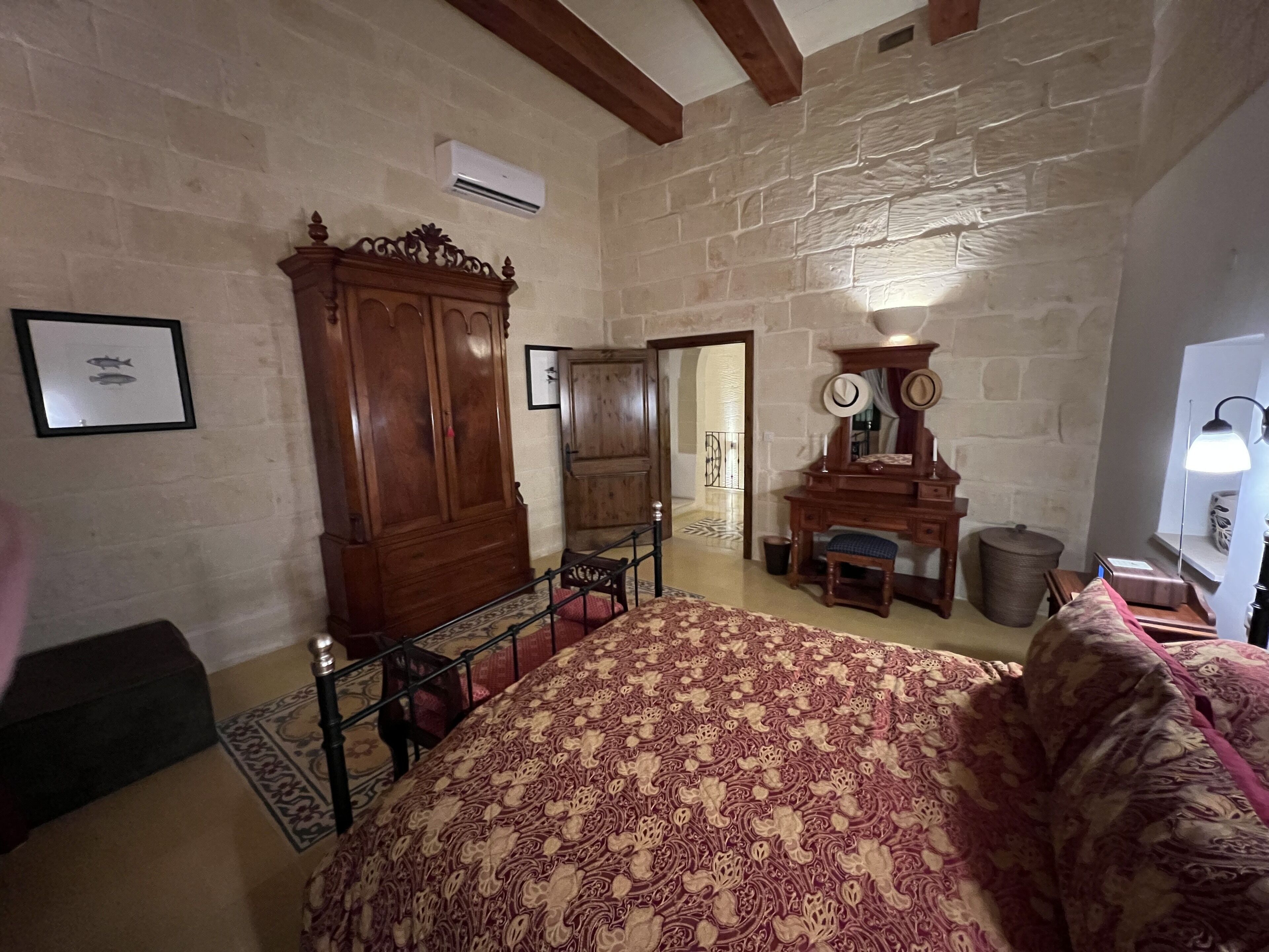 Ein Schlafzimmer mit einem großen Bett, einem Holzschrank und einer Kommode. Über der Kommode hängt ein Spiegel und auf dem Nachttisch steht eine Lampe. Die Wände sind aus Stein und die Decke aus Holz.