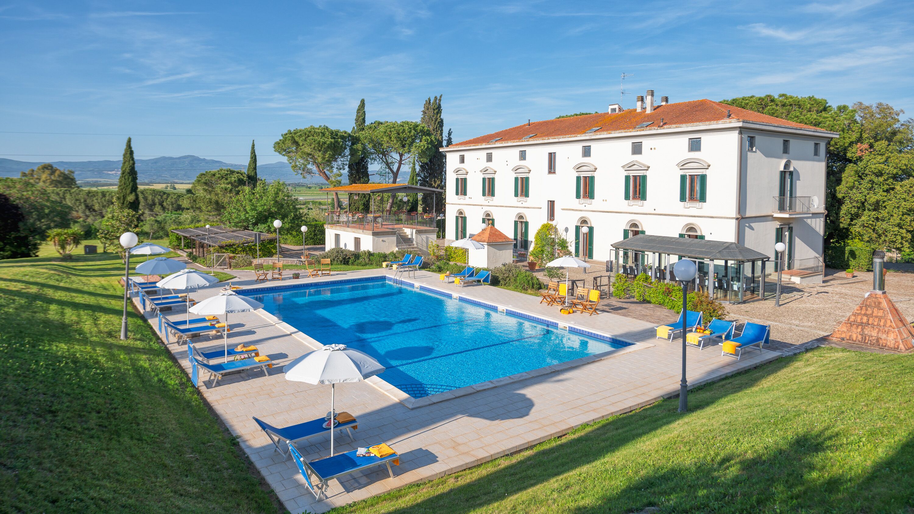A large swimming pool with sun loungers and umbrellas, surrounded by a well-maintained lawn and a building with a tiled roof.