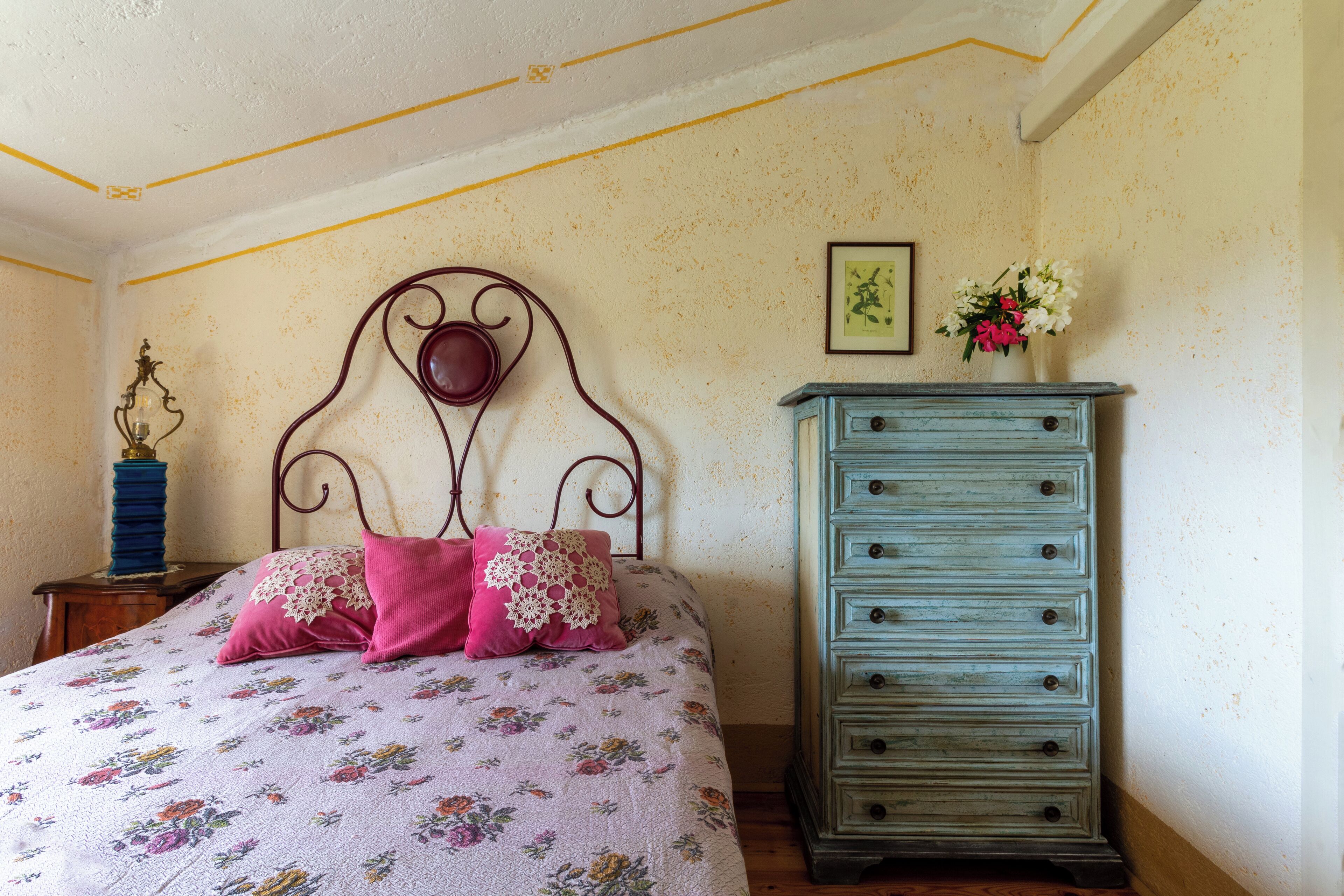 A bedroom with a floral bedspread, a metal headboard, a dresser, and a nightstand with a lamp.