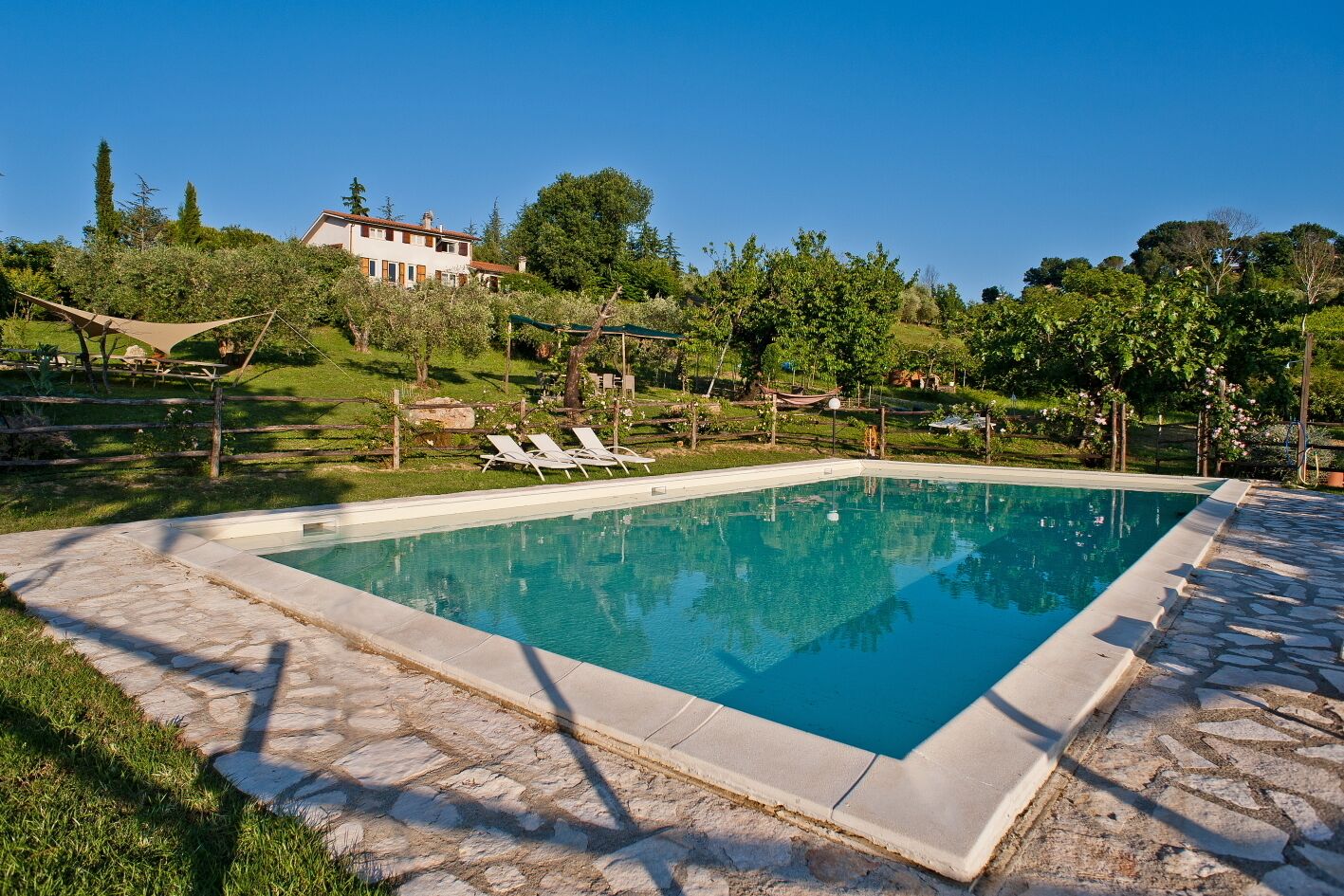 A rectangular swimming pool with a stone border, surrounded by a stone patio. There are lounge chairs and a wooden fence in the background.