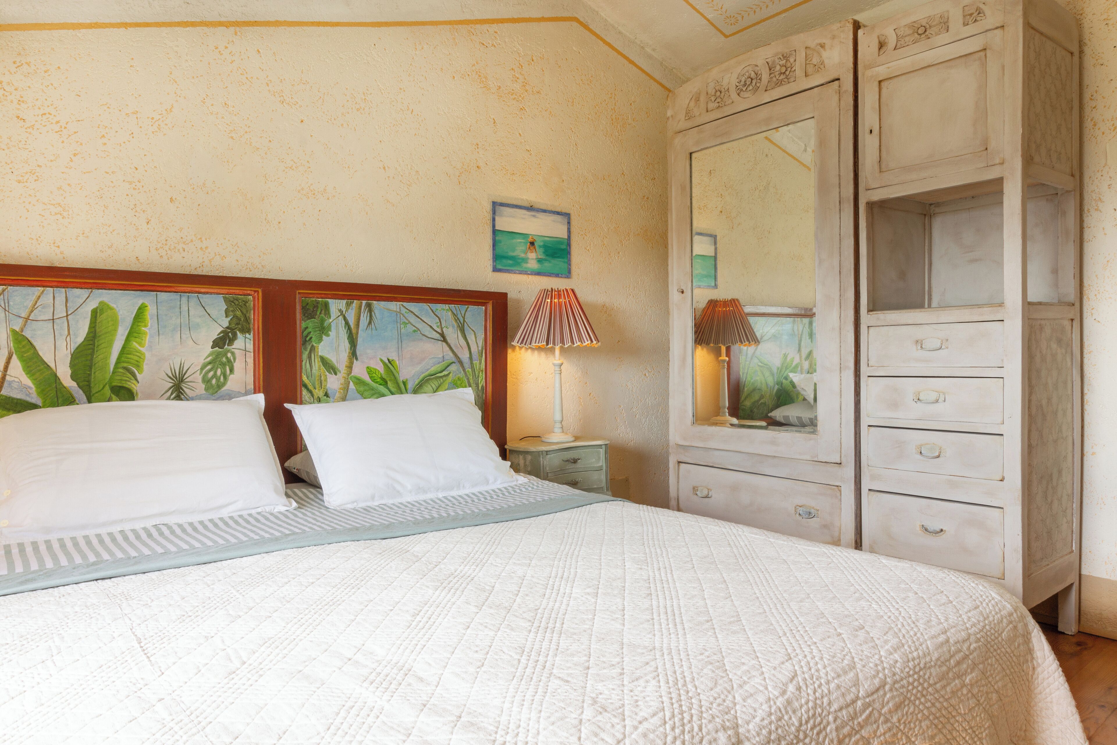 A bedroom with a bed, two bedside tables, a dresser with drawers, a mirror, and a painting on the wall.
