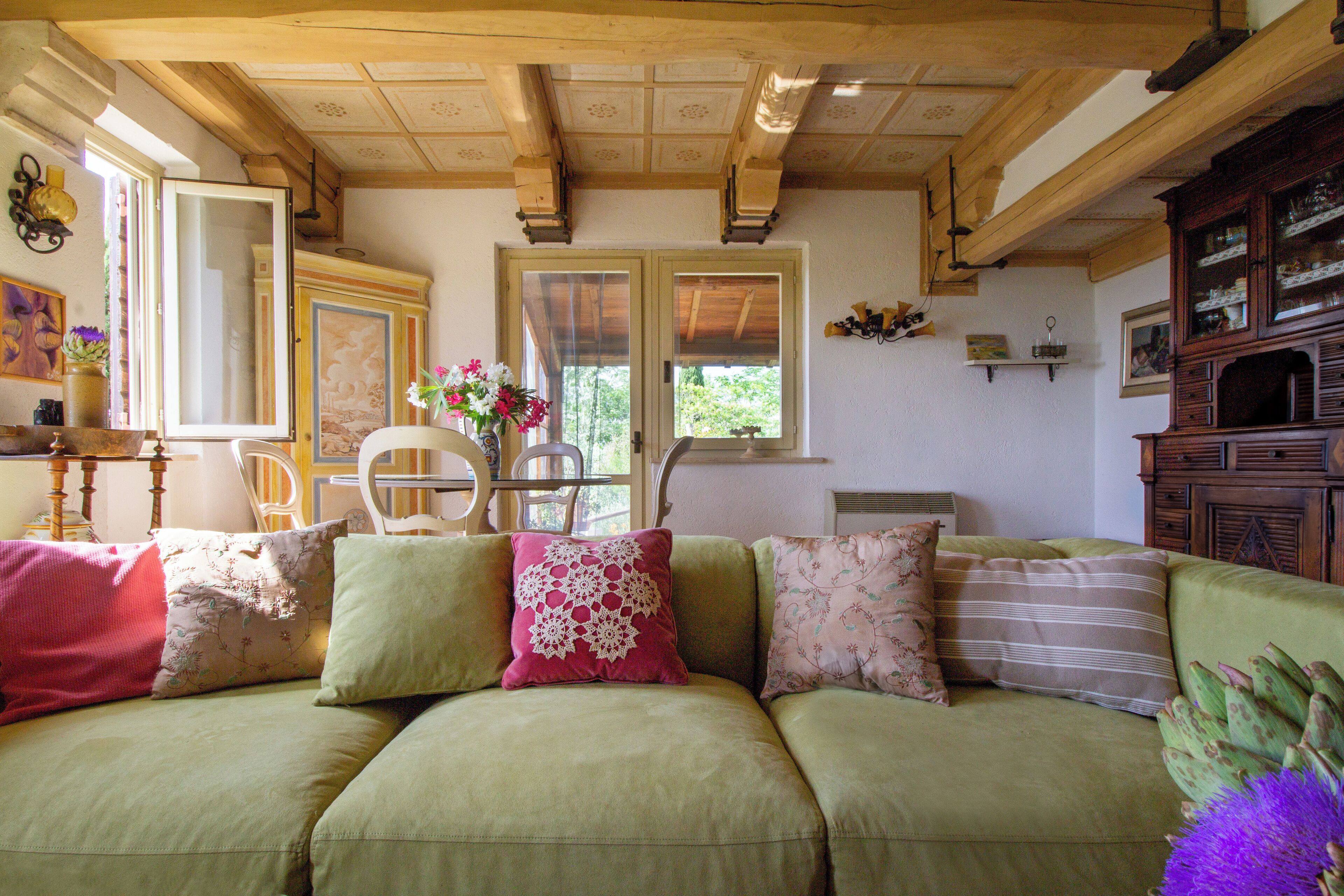 A cozy living room with a green sofa, a wooden ceiling, and a window with a view of greenery.