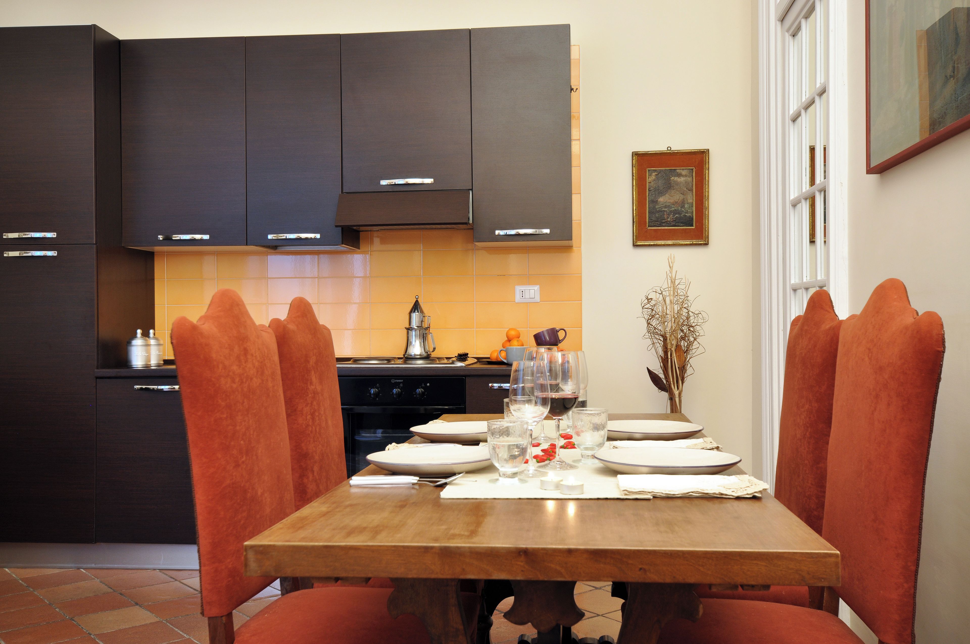 Un comedor con una mesa de madera dispuesta para cuatro personas, rodeada de sillas naranjas, en una cocina con armarios oscuros y un salpicadero de azulejos.