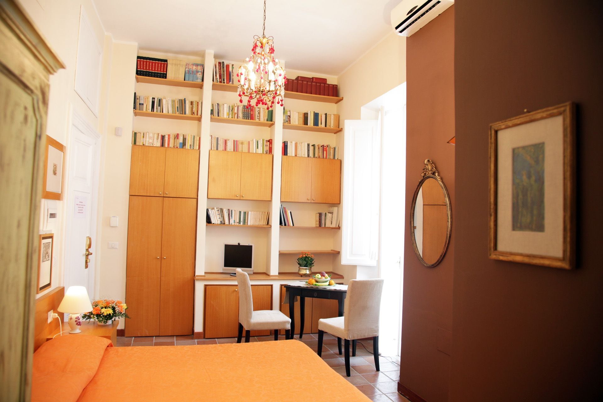 A hotel room with a bed, a desk with a chair, a bookshelf, a chandelier, and a framed picture on the wall.