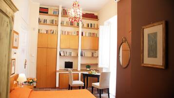 A hotel room with a bed, a desk with a chair, a bookshelf, a chandelier, and a framed picture on the wall.