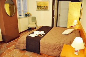 A hotel room with a bed, a chair, a bedside table with a lamp, a painting on the wall, and a window with a view.