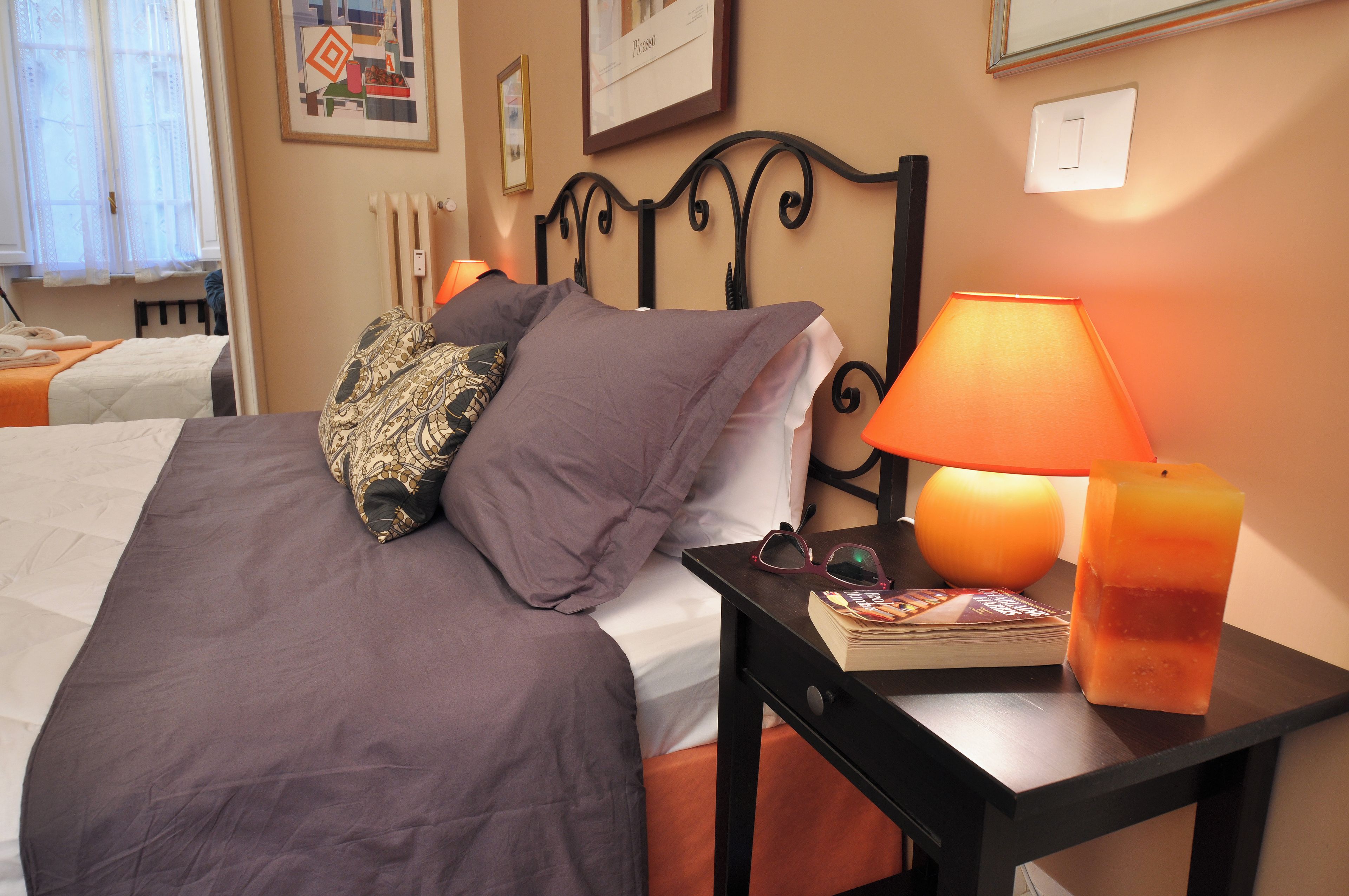 A neatly made bed with a patterned pillow, a bedside table with a book and glasses, and a lit orange lamp.