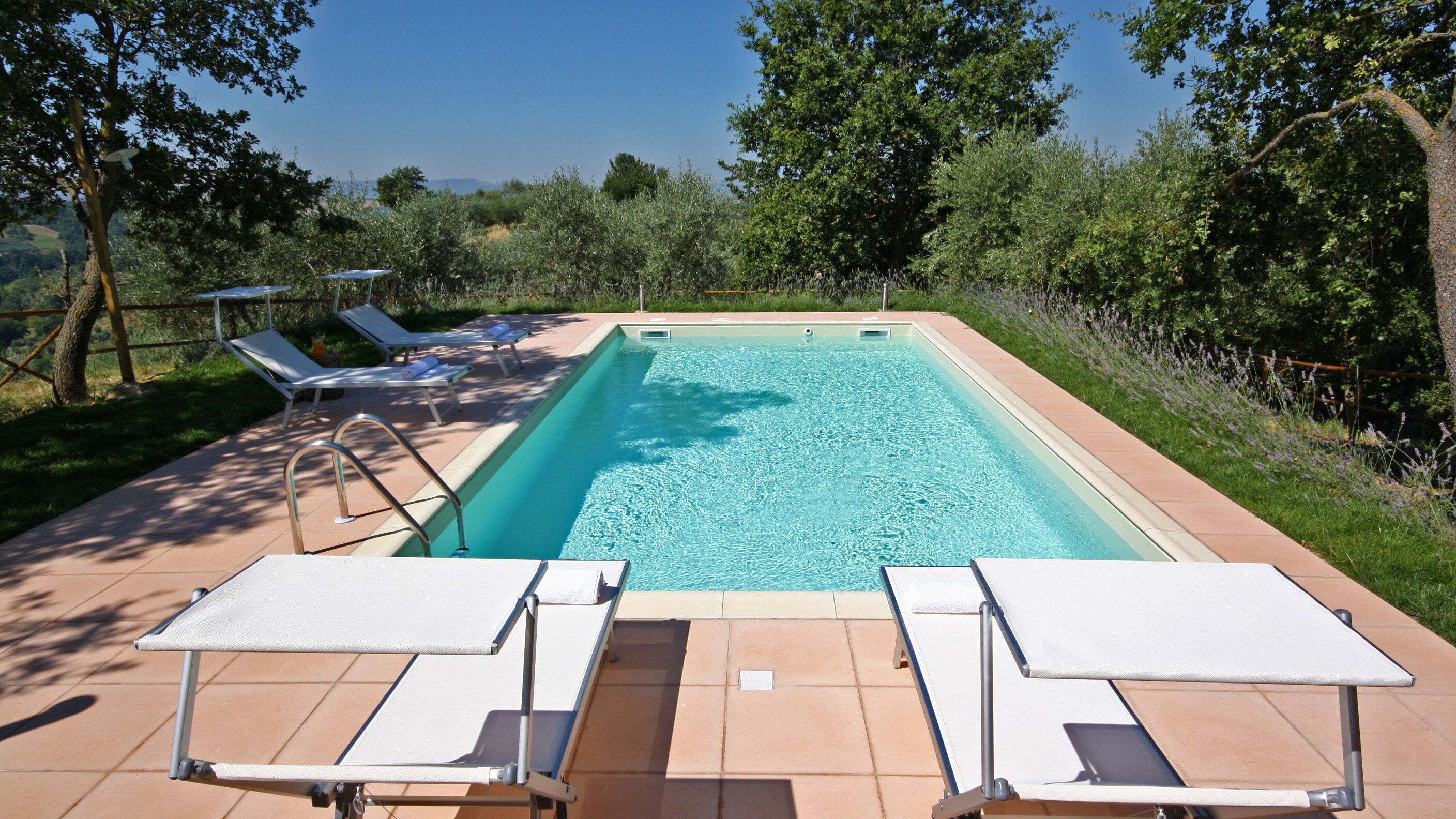 Une piscine rectangulaire à l’eau d’un bleu limpide, entourée d’une terrasse carrelée. On y trouve deux chaises longues blanches à structure métallique et un escalier en métal menant à la piscine. La piscine est située dans un espace aménagé avec des arbres et des arbustes, et un ciel dégagé se trouve au-dessus.