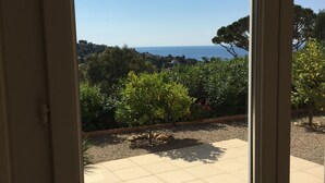 A view through a glass door to a patio with a tiled floor, surrounded by greenery and a distant view of the sea.