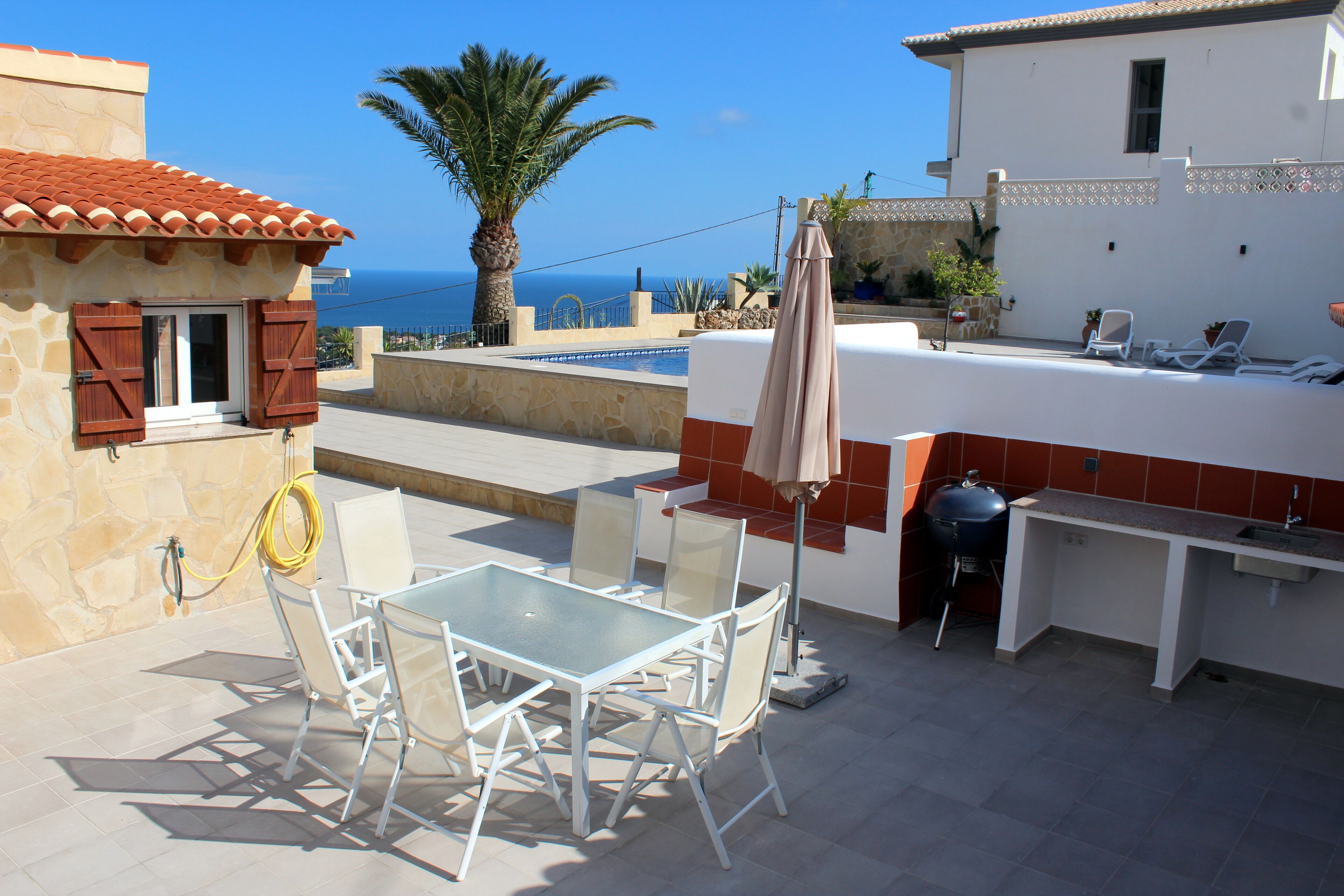 An outdoor patio with a table and chairs, a grill, and a view of the sea.