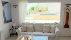A living room with a white sofa, a glass coffee table, a painting on the wall, a clock, and a view of a swimming pool. - Beautiful villa private pool stunning Panoramic Sea & Mountain Views in Moraira, (Moraira)