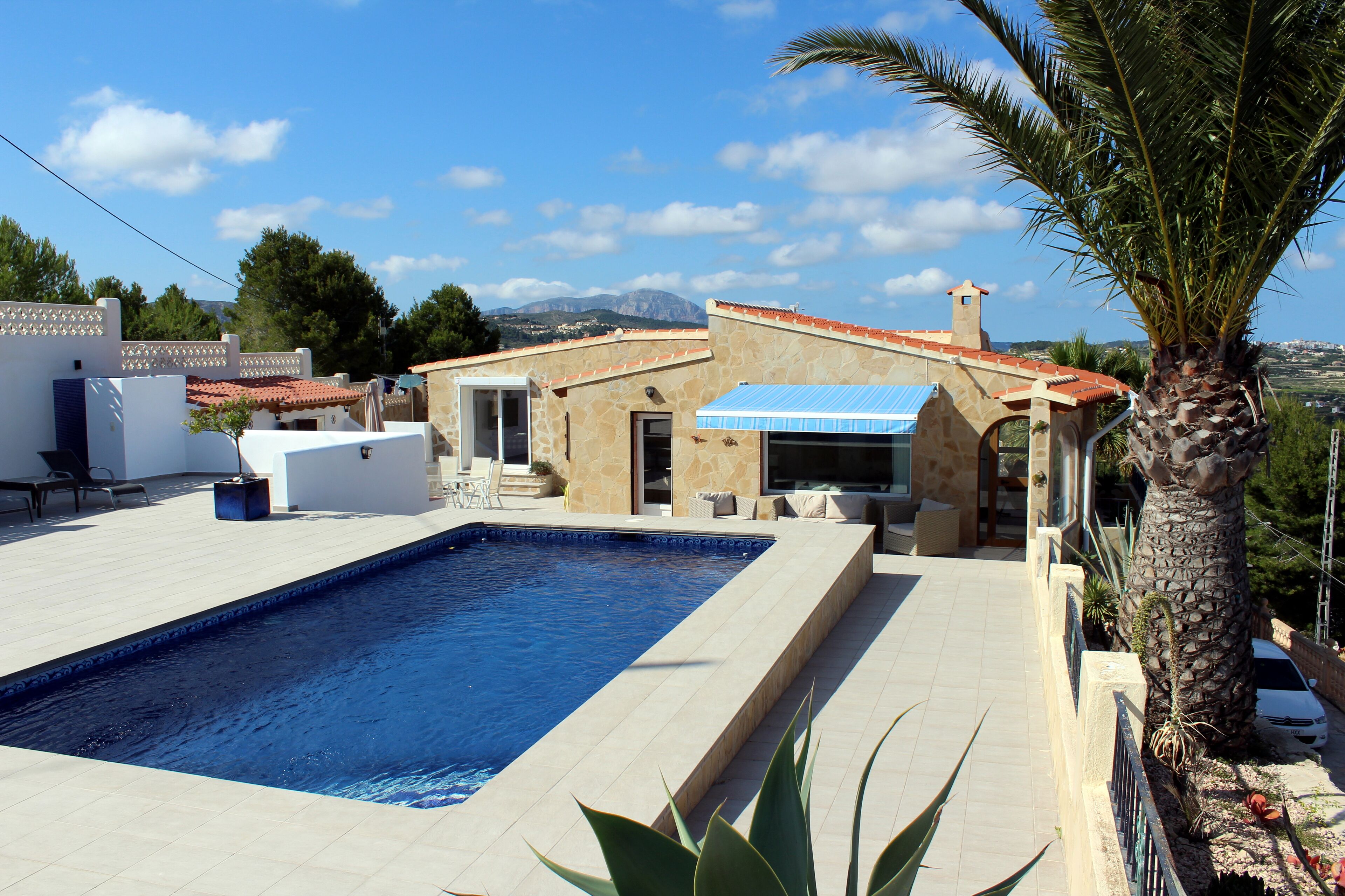 A swimming pool with a stone deck, a palm tree, and a building with a tiled roof.