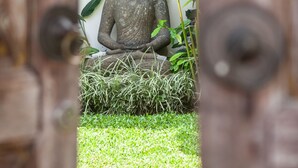 Eine Buddha-Statue in einem Garten, im Hintergrund ein Gebäude mit einem Reetdach.