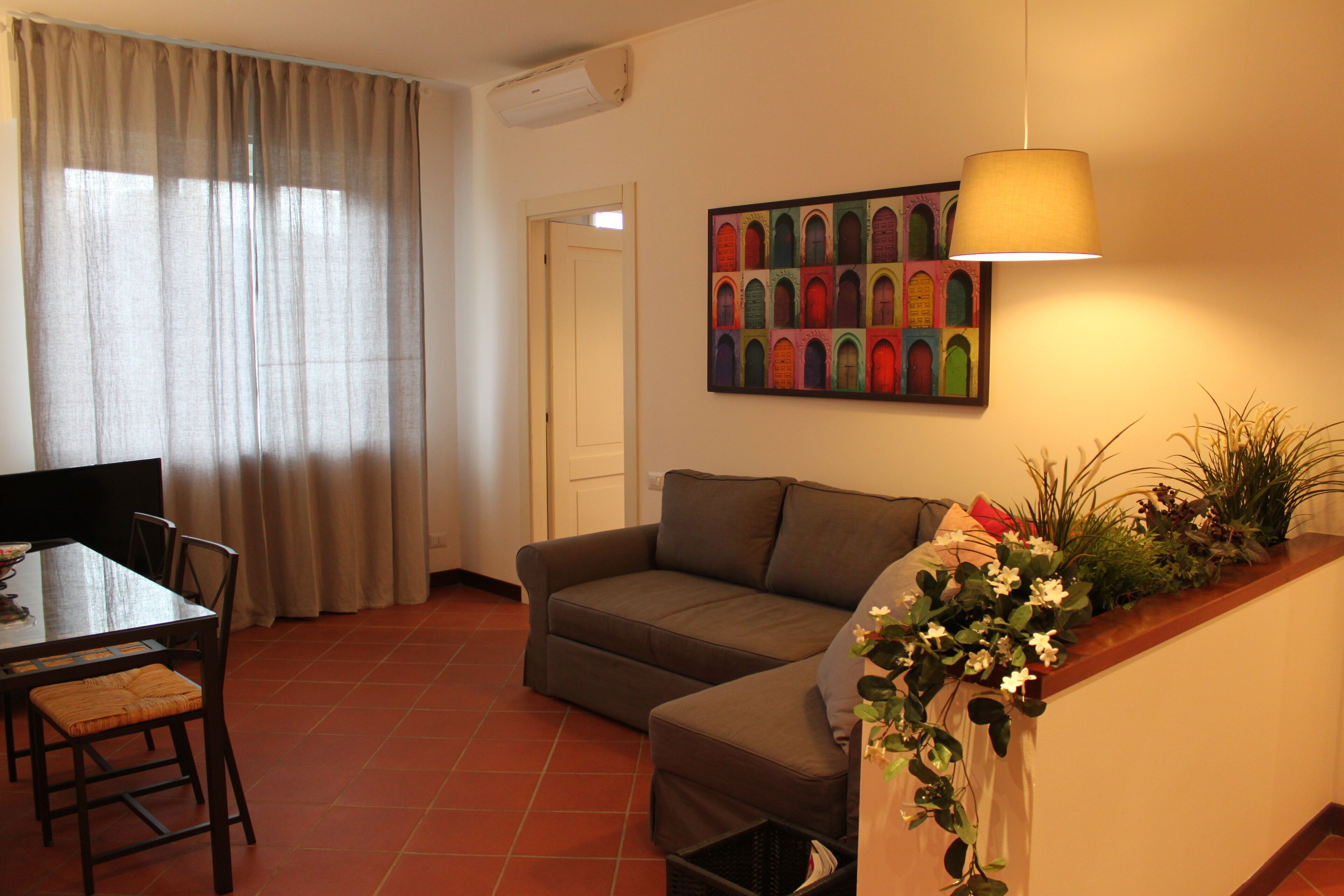 Ein Wohnzimmer mit einer Couch, einem Esstisch mit Stühlen, einem Fernseher, einem Fenster mit Vorhängen und einer Wandlampe.